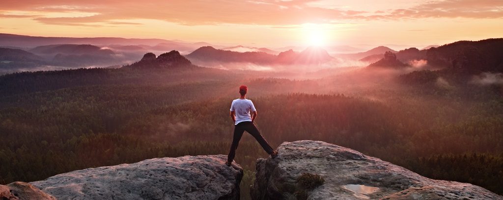 Photo or person with one foot on each of two rocks forming a gap in a canyon while facing a sunset or sunrise.