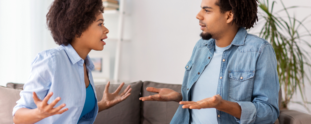 Couple arguing while sitting on sofa, facing each other with elbows bent and palms up.