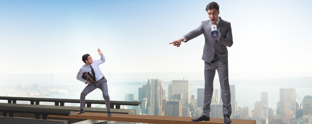 Photo of two people. One is larger than the other and points in shaming way. The smaller person puts a hand up with bent elbow to create a shield.